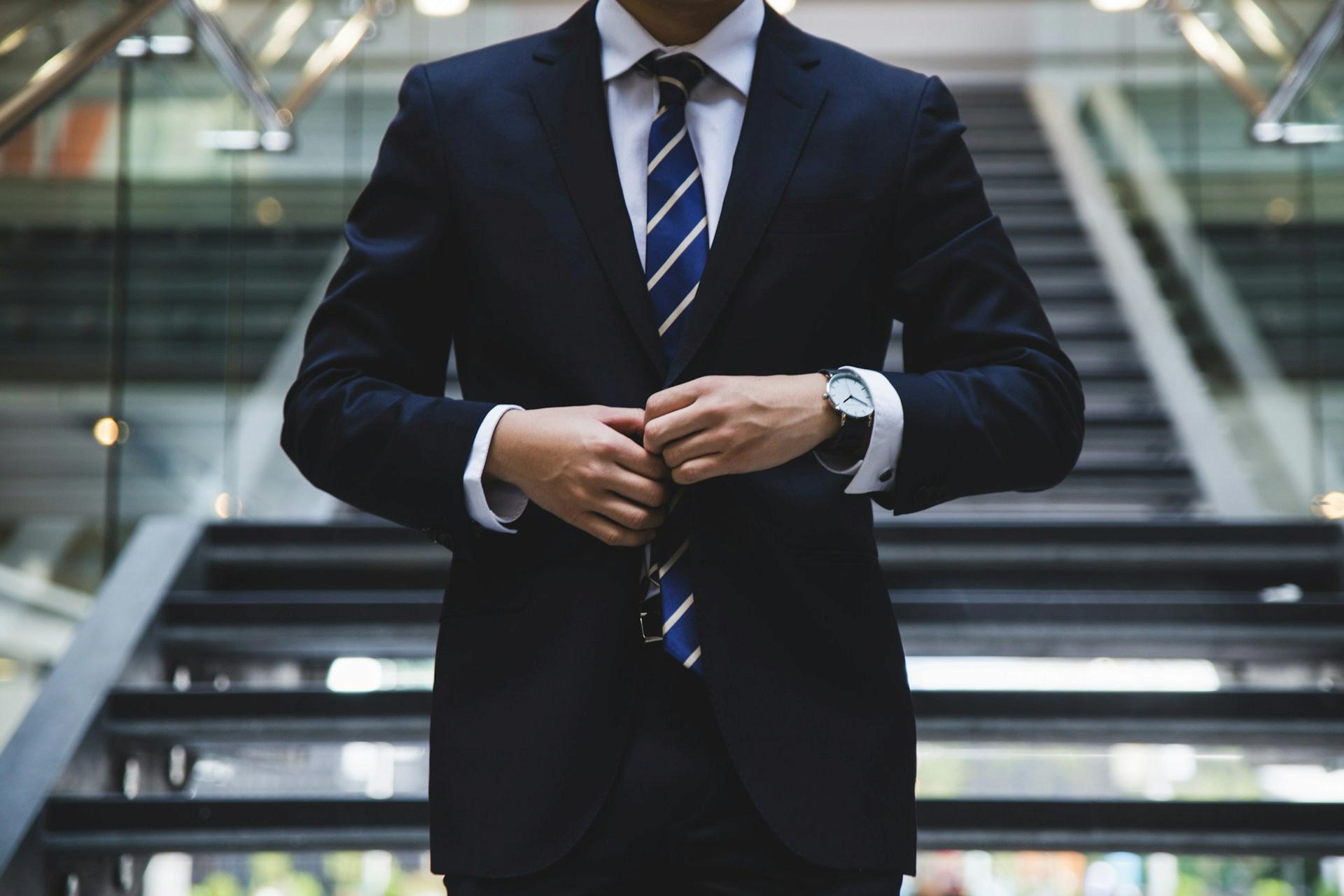A man in a dark suit fastens his jacket in front of a modern staircase.