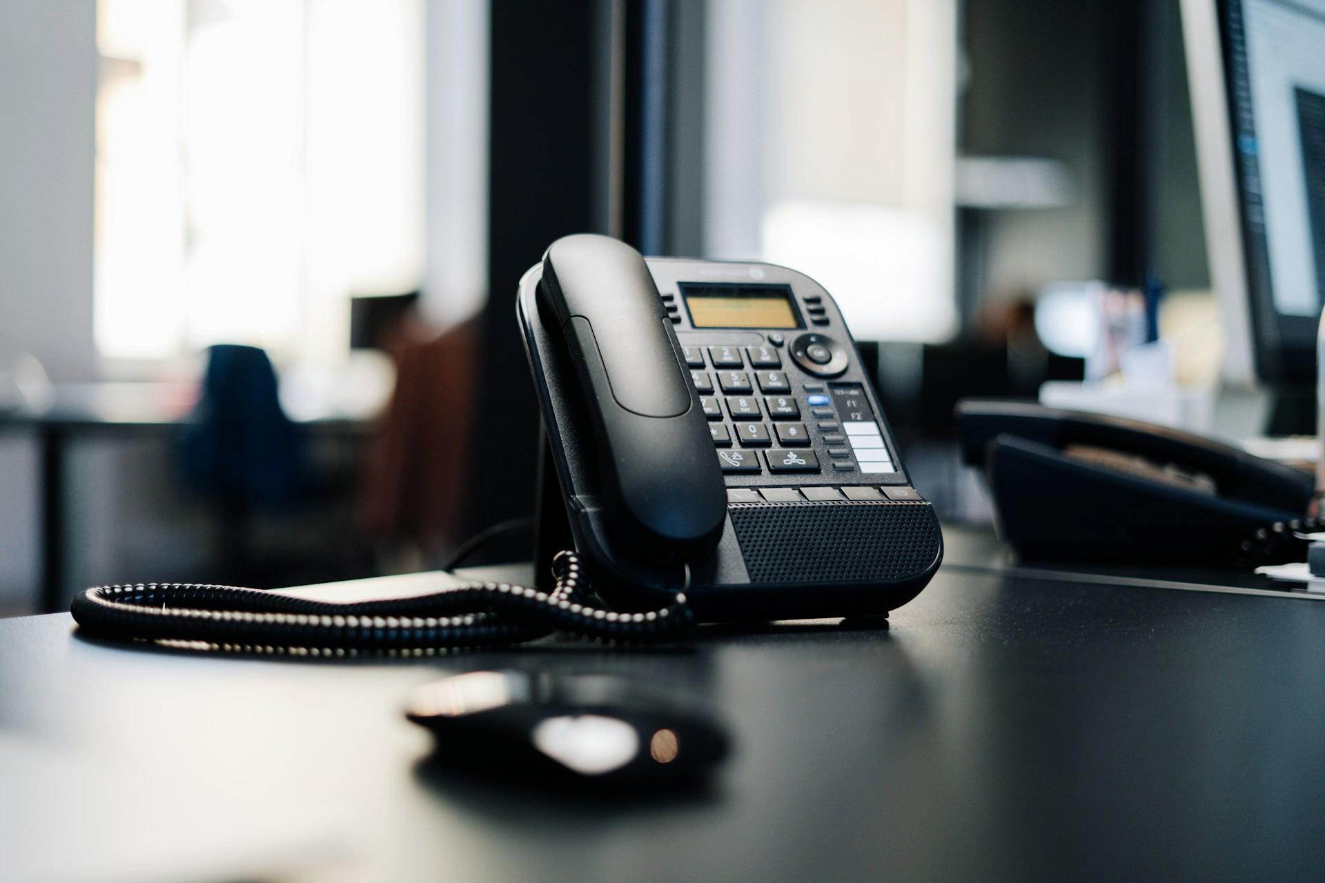 A black office desk phone is placed on a dark desk.