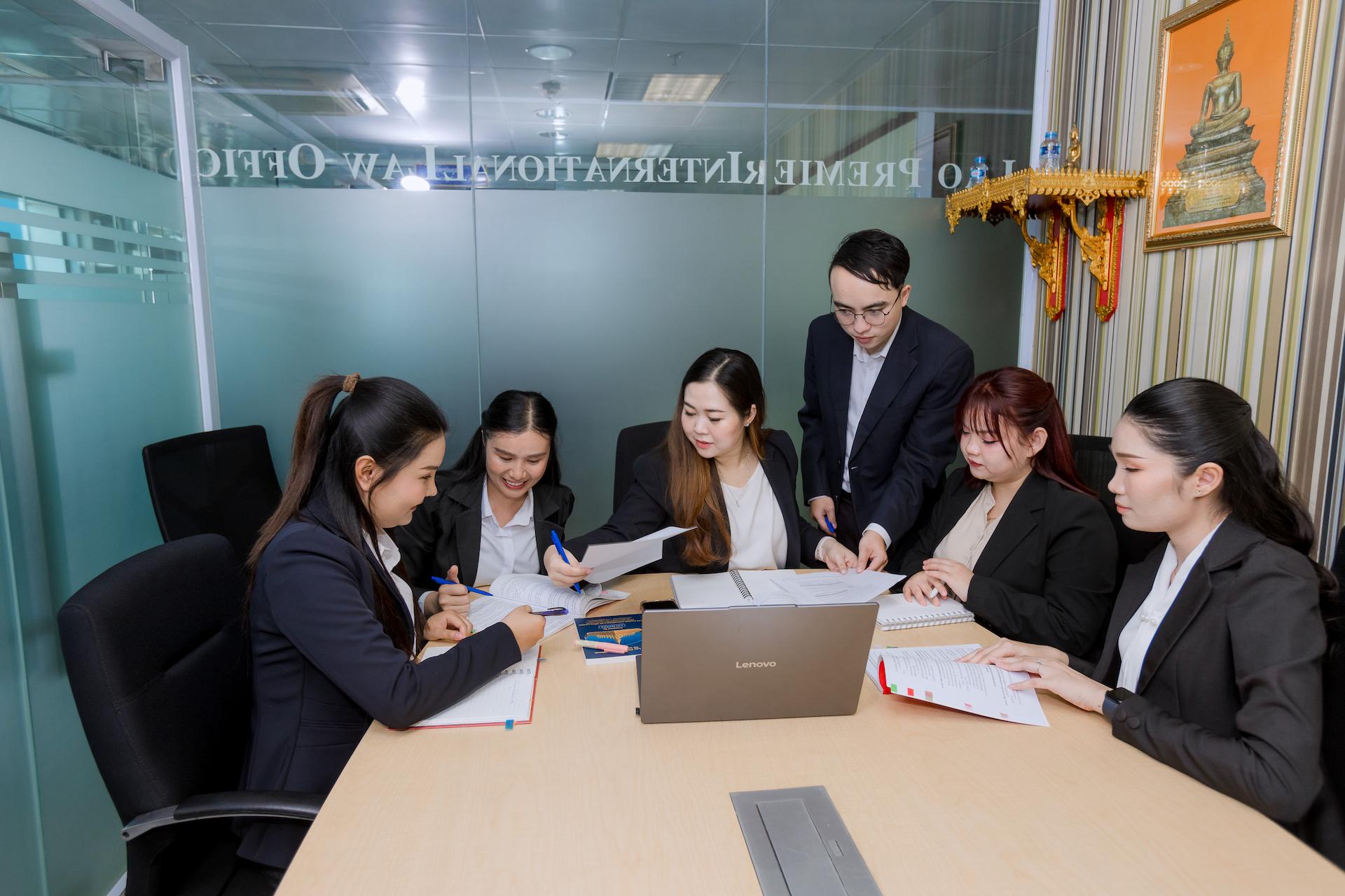 A group of people are gathered around a table in an office, looking at documents and a laptop.
