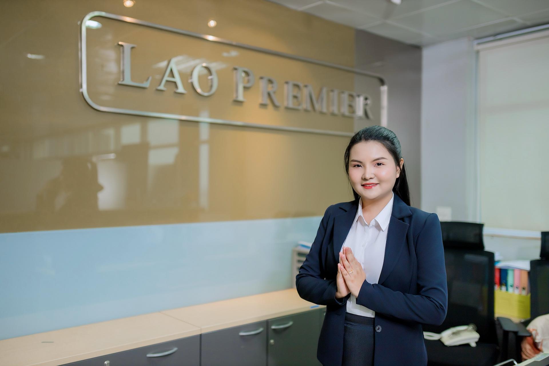 A young woman in a suit smiles and clasps her hands in front of her chest, standing in an office. Behind her is a wall with the words - LAO PREMIER - in large letters.