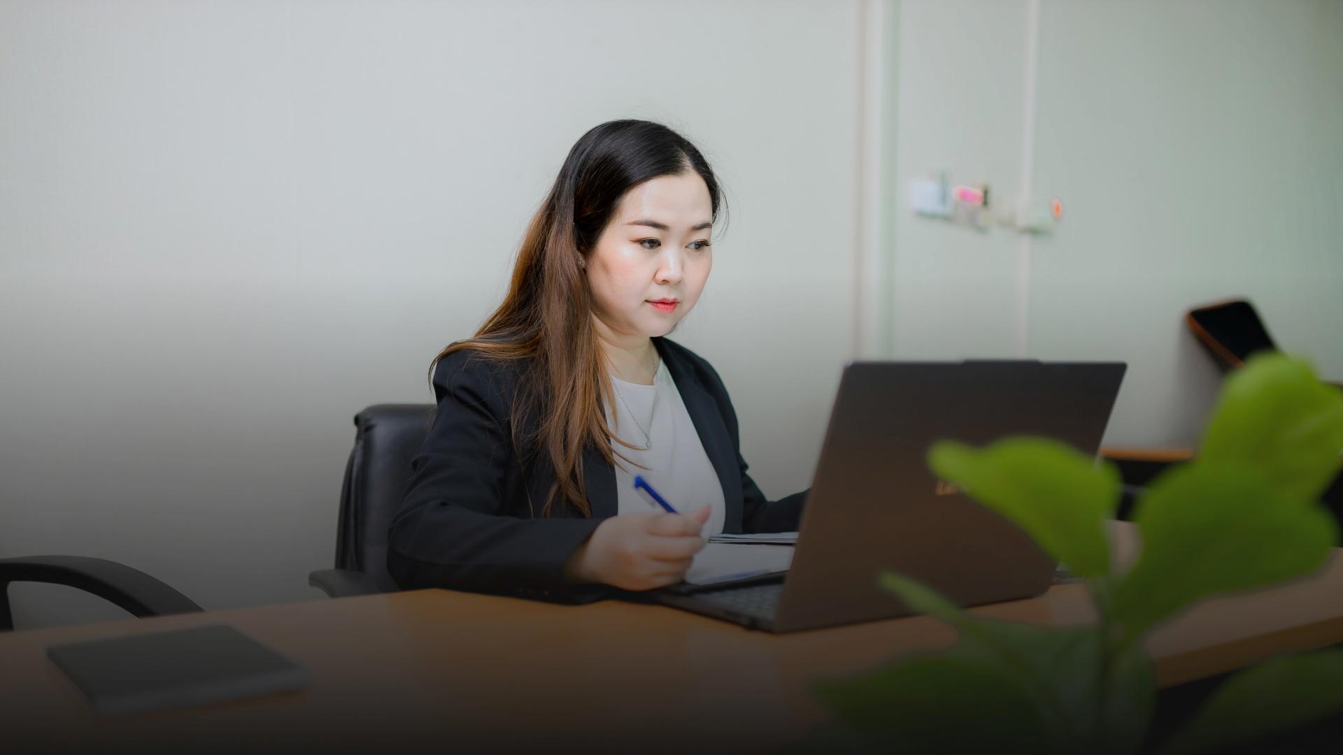 A woman in a suit is working on her laptop at a desk, holding a pen and looking at the screen. 