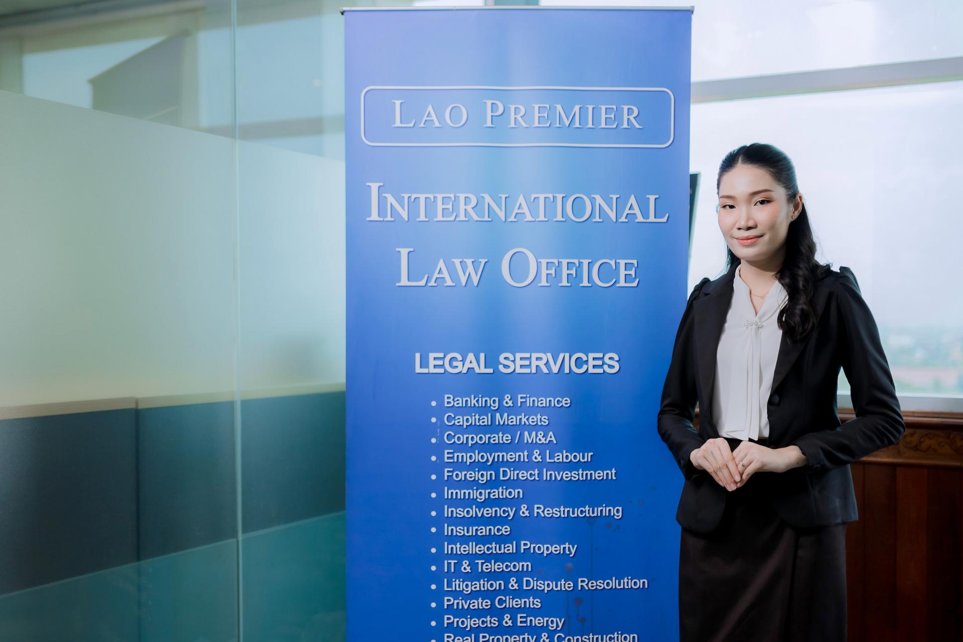 A woman stands in front of a blue banner that advertises 'LAO PREMIER INTERNATIONAL LAW OFFICE' and lists various legal services.